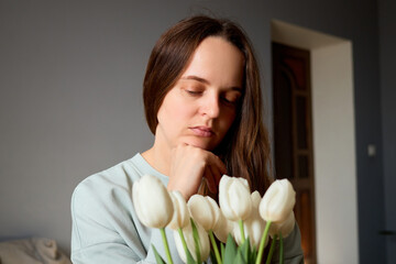 A woman is deep in thought, resting her chin on her hand, as she gazes at a bouquet of white...