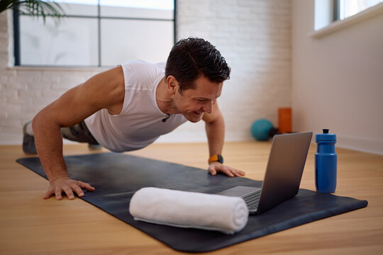 Happy athletic man doing push-ups while using laptop and working out at home.