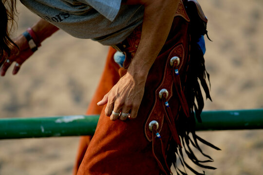 Cowboy wearing fringed leather chaps leaning over a green fence outdoors. Brandenburg, Germany