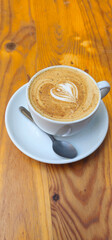 A white ceramic cup with a saucer holds a cappuccino with heart-shaped latte art; a teaspoon rests on the saucer; the wooden table has visible knots and grain;