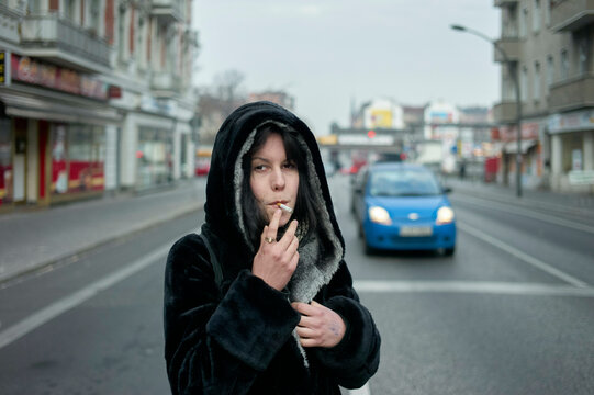 Woman in a black coat smokes on an urban street with a blue car approaching. Berlin, Germany