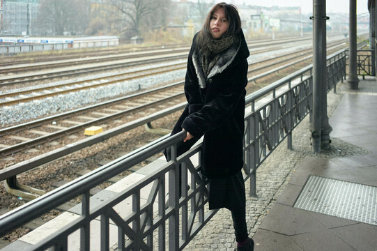 Woman in a dark coat stands by a railing at an empty train station on a cloudy day. Berlin, Germany