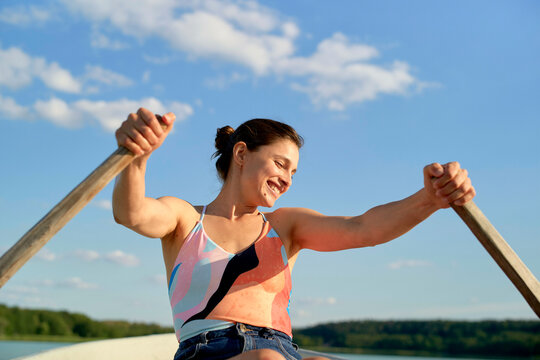 Woman rowing a boat on a sunny day, smiling with a colorful tank top, against a clear sky. Brandenburg, Germany