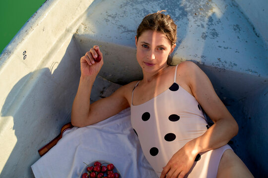 Woman in polka dot swimsuit lounging in boat with cherries and sunlight casting shadows. Brandenburg, Germany