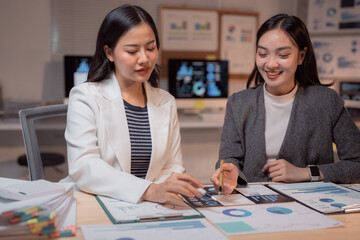 Fototapeta premium Two young Asian businesswomen analyzing financial charts and graphs, working together on a project in a modern office, demonstrating teamwork and dedication