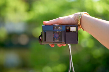 Female hand holding a vintage compact camera taking a selfie against a blurred greenery background.