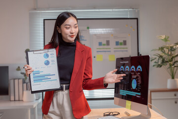 Young professional woman presenting marketing data analysis during a business meeting, using a clipboard and a desktop computer, standing in her office