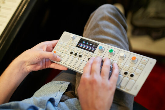 Hands playing a compact electronic synthesizer with colorful knobs on a person's lap in a recording studio.
