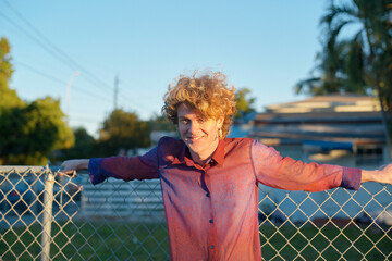 Smiling person with curly hair wearing a red shirt leans against a chain-link fence outdoors. Miami, FL, USA