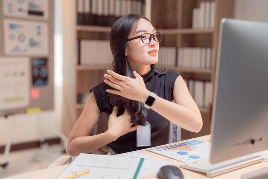 Young businesswoman adjusting her hair while working on computer at her desk in a modern office, she is wearing glasses and a smartwatch and looking away from the screen - Powered by Adobe