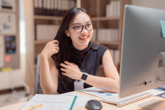 Young happy asian businesswoman working from her modern office using computer, she's smiling and playing with her hair while looking at the monitor
