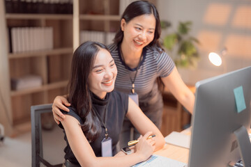 Two Asian businesswomen collaborating at a modern office desk, working together on a desktop computer while smiling, embodying teamwork and partnership in a professional environment
