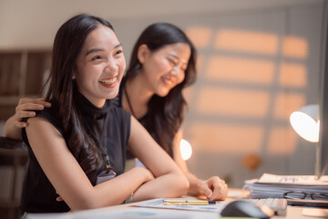 Two young asian businesswomen are working together in a modern office, one of them is putting a hand on the shoulder of her colleague, they are smiling and looking away