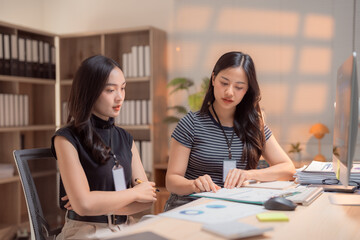 Two young asian businesswomen wearing name tags are sitting at desk and analyzing financial graph, discussing marketing strategy and making notes while working on new project in modern office