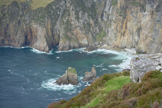 Rugged cliffs and waves crashing against rocks on a scenic coastline with green hills. Ireland