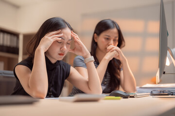 Two Asian businesswomen experiencing headaches and stress while working at their desks in the...