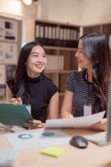 Two young businesswomen are smiling and discussing marketing strategy using data charts and graphs, working together on a project in modern office