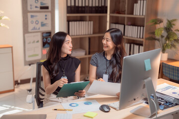 Two asian businesswomen are discussing and working together using computer and documents in a modern office, analyzing financial charts and statistics, collaborating on a project and sharing ideas