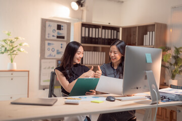 Two happy Asian businesswomen smiling and giving thumbs up while reviewing documents together in a modern office, showcasing collaboration and teamwork in a professional environment