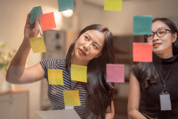 Two Asian businesswomen collaborating at the office, using colorful sticky notes on a glass wall to brainstorm and plan innovative strategies for their business growth and success