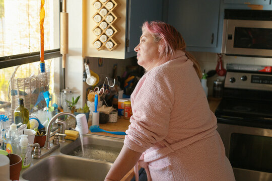 Woman in a pink robe gazing out the kitchen window while doing dishes. California, USA