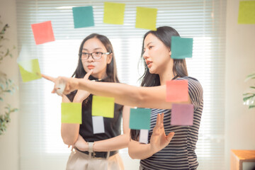 Two asian businesswomen are discussing and planning strategy using sticky notes on a glass wall in a modern office, sharing ideas and collaborating on a project