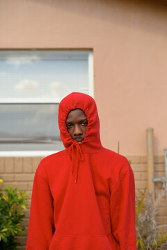 Young man in a red hoodie stands in front of a window on a beige wall with plants nearby. Miami, Florida
