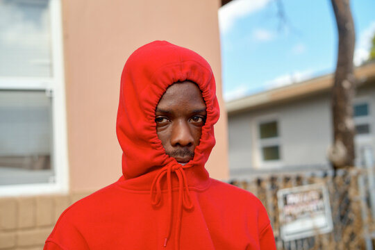 Young man in a red hoodie stands outdoors in a suburbian area, face mostly covered, looking at the camera. Miami, Florida