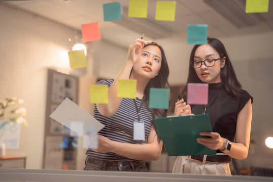 Two asian businesswomen sharing ideas using colorful sticky notes and writing on clipboard, working together on new project plan in modern office at night