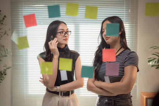 Two asian businesswomen are brainstorming and planning their strategy using colorful sticky notes on a glass wall in a modern office, discussing and sharing ideas for their next project