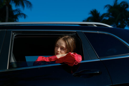 Woman in red shirt leaning out of car window under blue sky with palm trees in the background. Miami, FL, USA