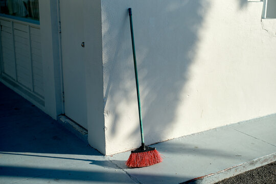 A red-bristled broom leans against a white wall, casting a shadow on a cement sidewalk. Miami, FL, USA