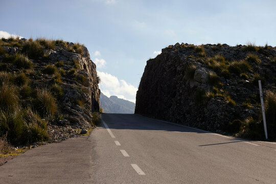 A rural road passes through two rocky hills under a clear blue sky with scattered clouds. Mallorca, Spain