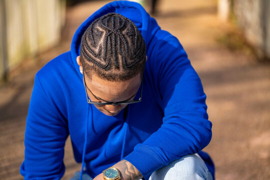 Young man in a blue hoodie kneels on a path, showcasing intricate braided hairstyle. Arnhem, Netherlands