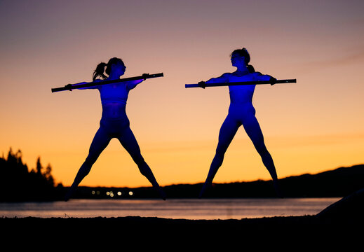 Two people practice acrobatic movements with poles at sunset by a calm waterfront. Bainbridge Island, WA, USA