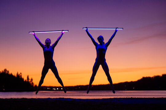 Two pwomen perform a jump with glowing sticks against a vibrant sunset backdrop by a lake. Bainbridge Island, WA, USA