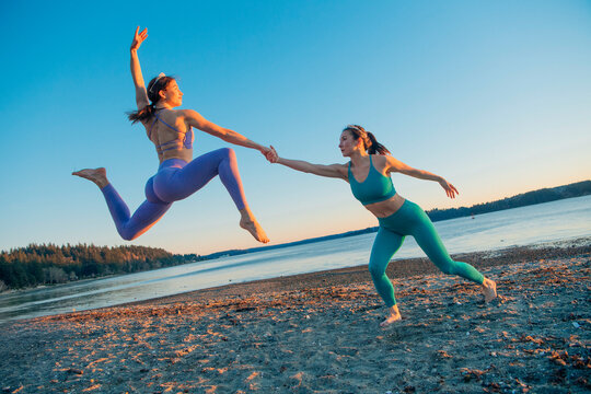 Two women in athletic wear performing a dynamic dance move on a beach at sunset. Bainbridge Island, WA, USA