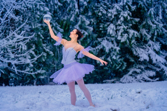 Ballerina in a lavender tutu dances in a snowy forest holding a clear orb above her head. WA, USA