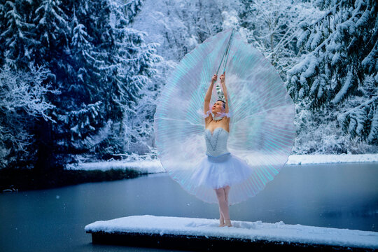 Ballerina in white tutu poses gracefully on snow-covered pier by a frozen forest lake. WA, USA