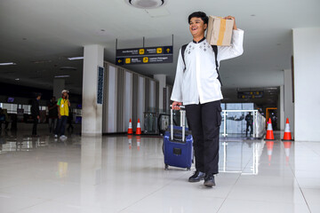 Young Muslim Man Carrying Cardboard Box and Pulling Suitcase at Terminal, Ready for Mudik during Eid Mubarak