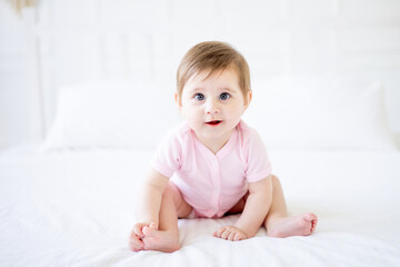 Fototapeta premium a six-month-old baby is sitting on a white cotton bed at home, a cute little baby girl in a pink bodysuit is smiling and rejoicing, close-up