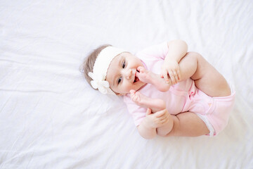 a six-month-old baby is playing with his legs lying on the back on a white cotton bed at home, a cute little baby girl in a pink bodysuit, close-up, top view