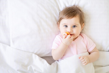baby sucking a pacifier lying on a white cotton bed under a blanket on the bed at home and smiling,...