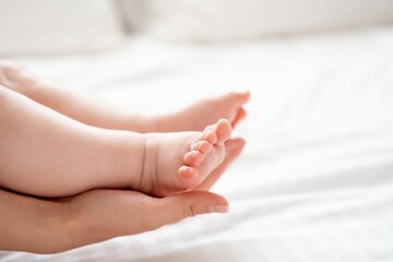 Mom's hand holds the small legs of her newborn baby on the white background of the bed, space for text