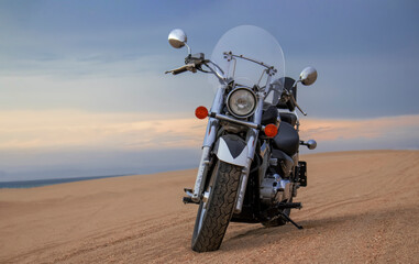 A lone motorcycle stands on the sandy surface of the Namib Desert against a dramatic cloudy sky. The sunset lighting highlights the contrast between nature and machine.