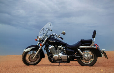 Obraz premium A lone motorcycle stands on the sandy surface of the Namib Desert against a dramatic cloudy sky. The sunset lighting highlights the contrast between nature and machine.