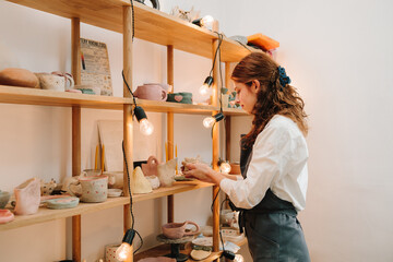 Woman in black apron arranges handmade pottery earthenware on wooden shelves decorated with cozy lights in the ceramic studio.