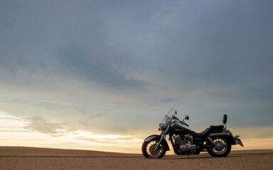 Fototapeta premium A lone motorcycle stands on the sandy surface of the Namib Desert against a dramatic cloudy sky. The sunset lighting highlights the contrast between nature and machine.