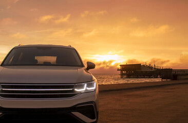Obraz premium A close-up of a gray car parked on the seashore at sunset. The soft light highlights its silhouette, creating a sense of solitude and freedom against the calm waves and vibrant sky.