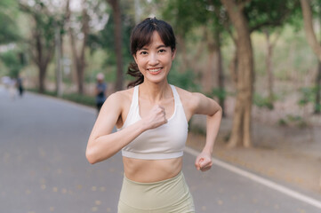 Asian woman jogging outdoors in park, wearing white sports bra and light green leggings, smiling confidently while enjoying her workout. Healthy and active lifestyle concept
